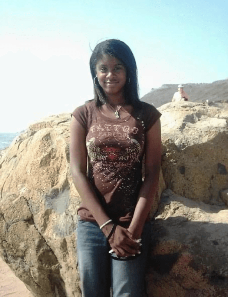 Ashleigh Warren, a person stands on a rock by the water, wearing a brown top and jeans, with a scenic background of hills and sky.