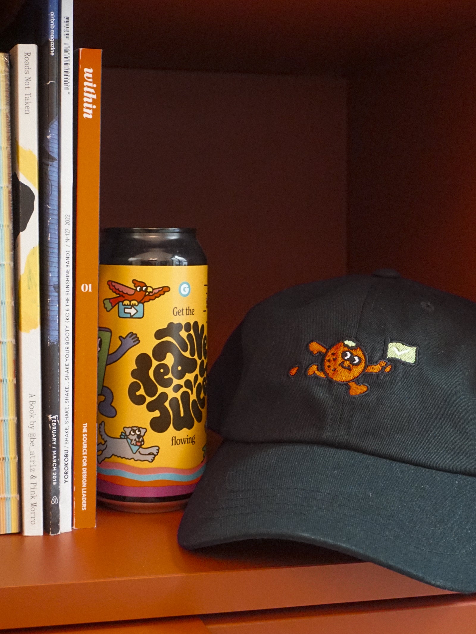 Black cap with an orange fruit design and a colorful can on an orange shelf with books