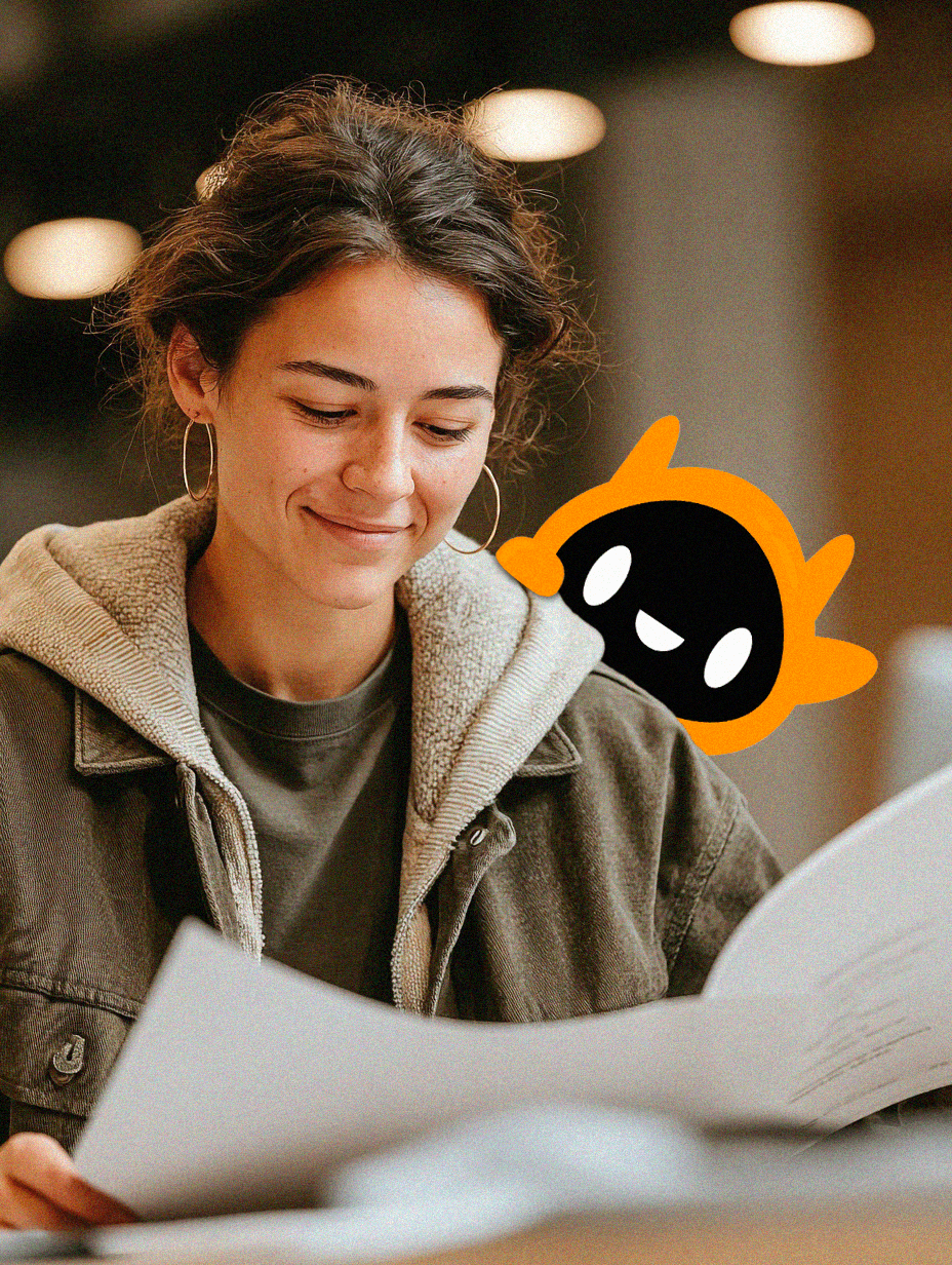 A young woman with her hair loosely tied back and wearing hoop earrings smiles gently while looking down at a sheet of paper she is holding. She is dressed in layered casual clothing, including a hoodie under a jacket. The background is softly lit with warm tones and blurred lights, suggesting an indoor setting such as a classroom, office, or café.