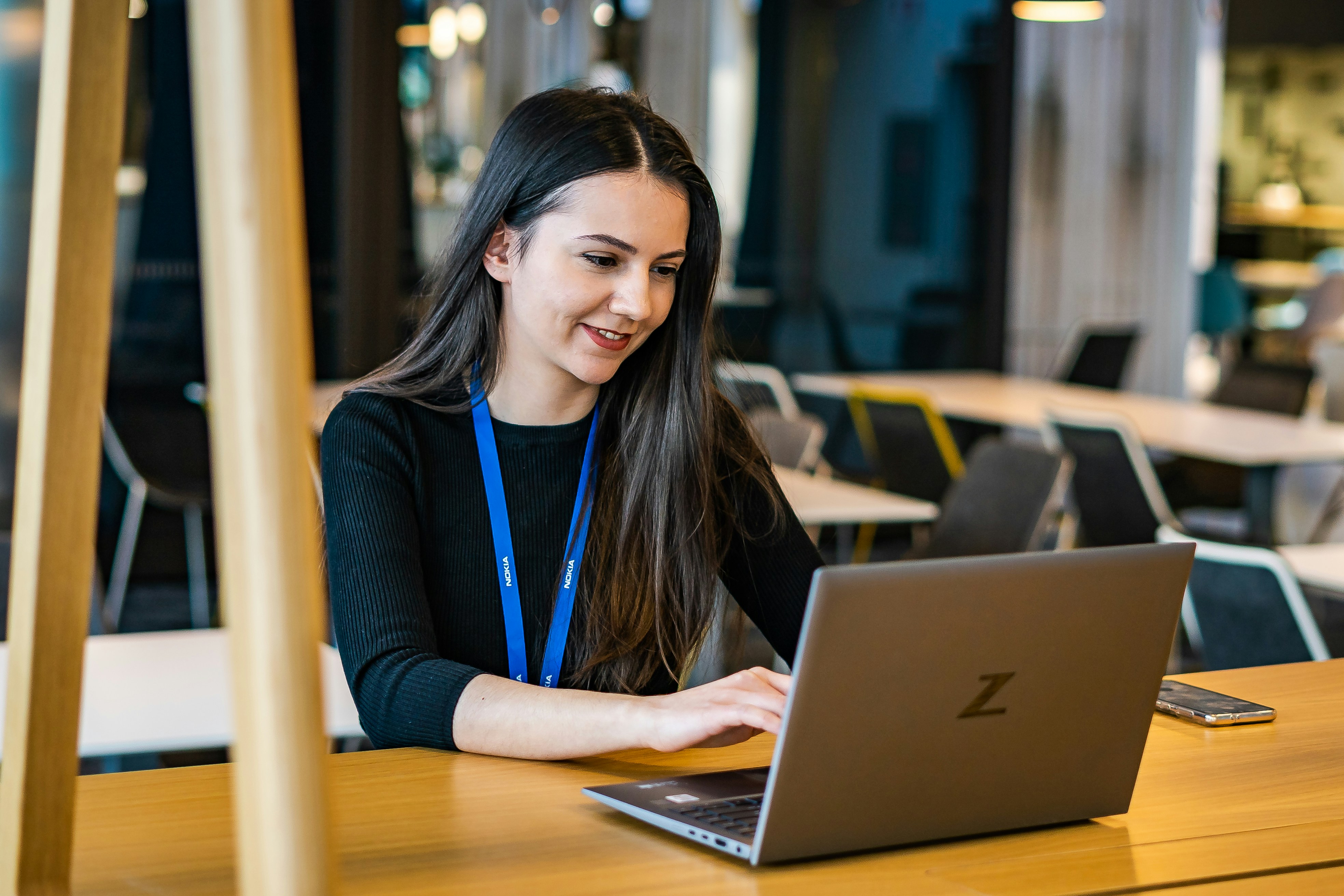 A smiling young woman looks down at a laptop, which is on a wooden desk. The background shows empty chairs and tables.