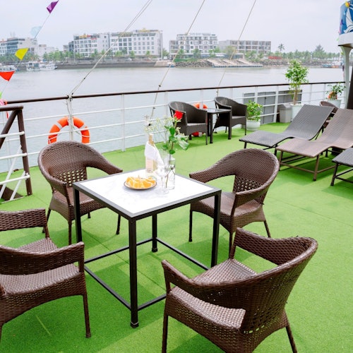 Udendørs siddeområde på en båd med kurvestole omkring et bord, liggestole og udsigt til vand og bygninger i baggrunden.