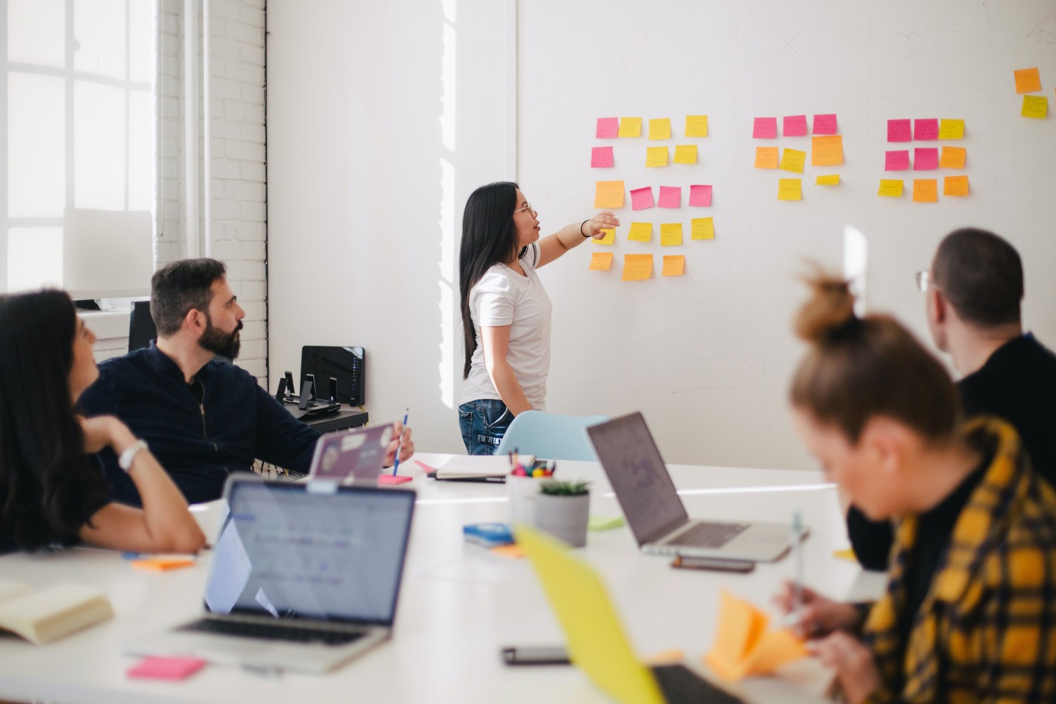 Group of five people in a conference room. One at the white board with sticky notes, the others seated around a conference table with their computers, pens, and sticky notes. Credit: Jason Goodman - Unsplash