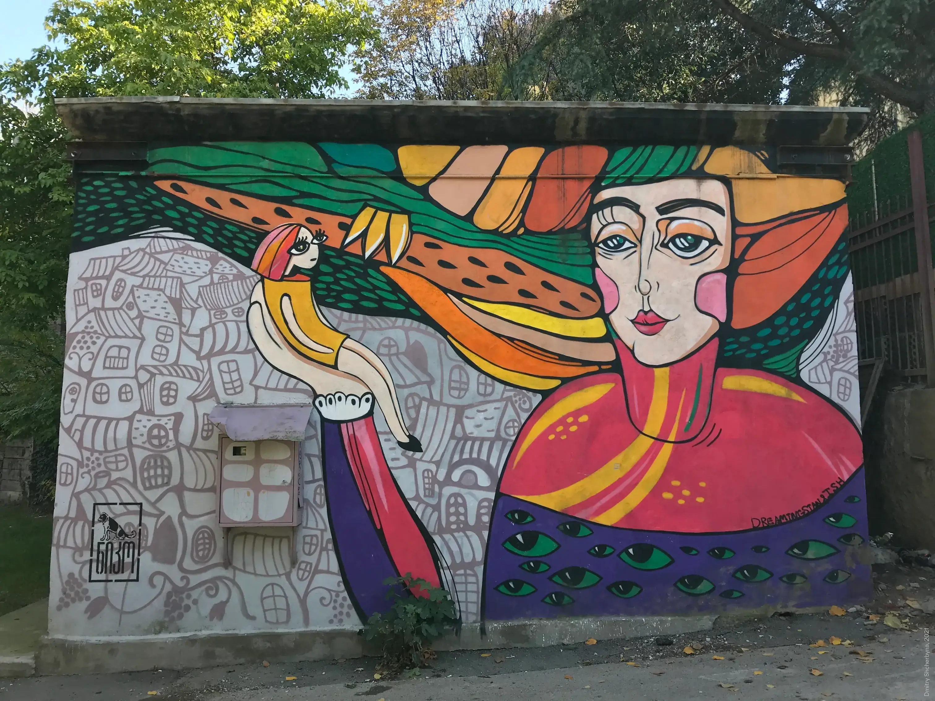 A colorful mural on a low concrete building depicts a large woman in a red and purple outfit with flowing, multicolored hair that stretches across the wall, cradling a small child figure. In the background, there are hand-drawn houses with watchful eyes.