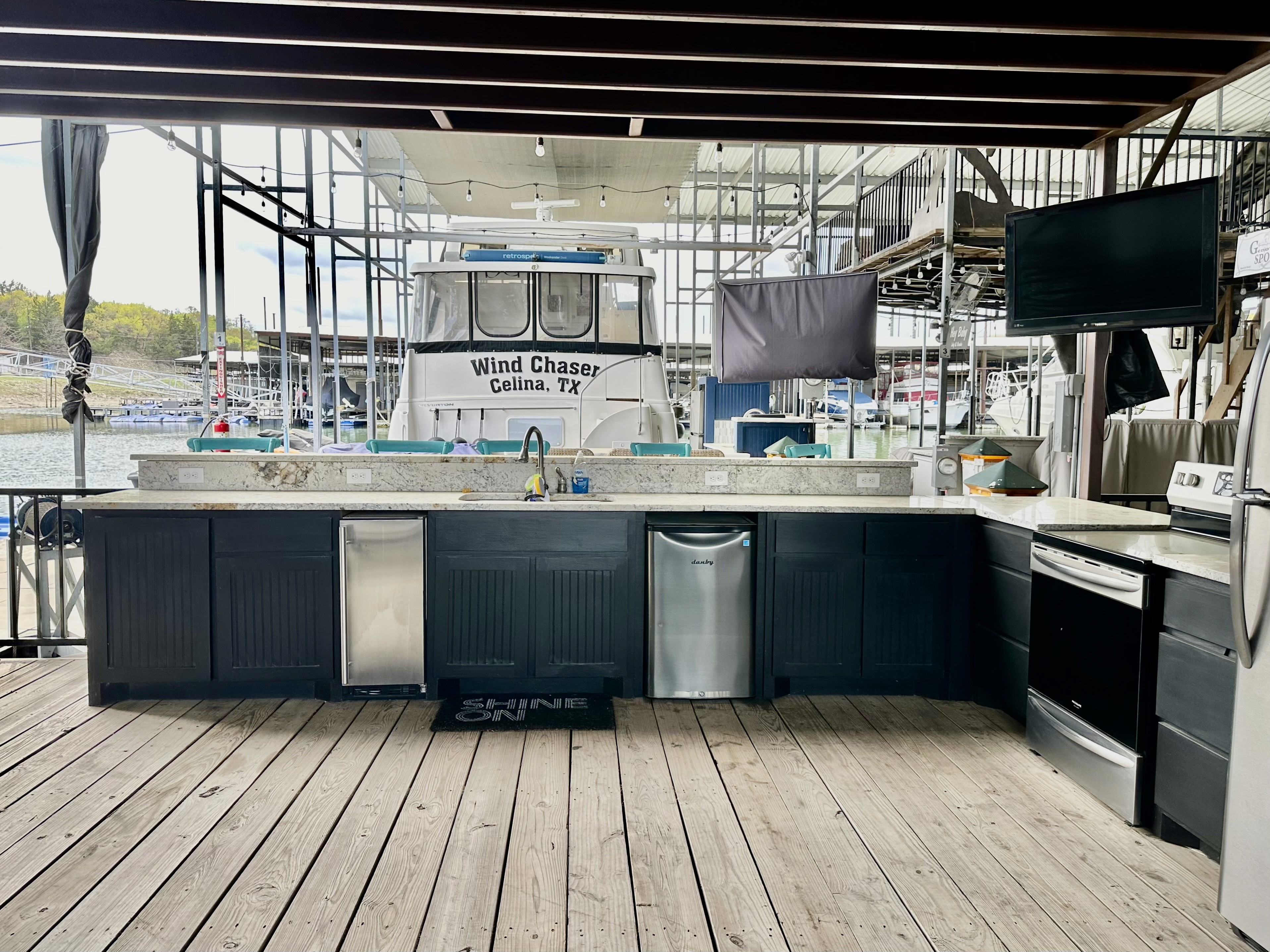 Outdoor dockside kitchen with a long granite countertop, stainless steel appliances including a dishwasher and refrigerator, and a view of the marina and docked boat in the background.