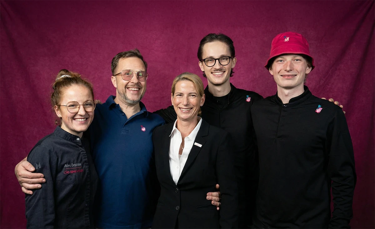 Three chefs in an open kitchen, one smiles holding a platter of desserts while the others prepare food.
