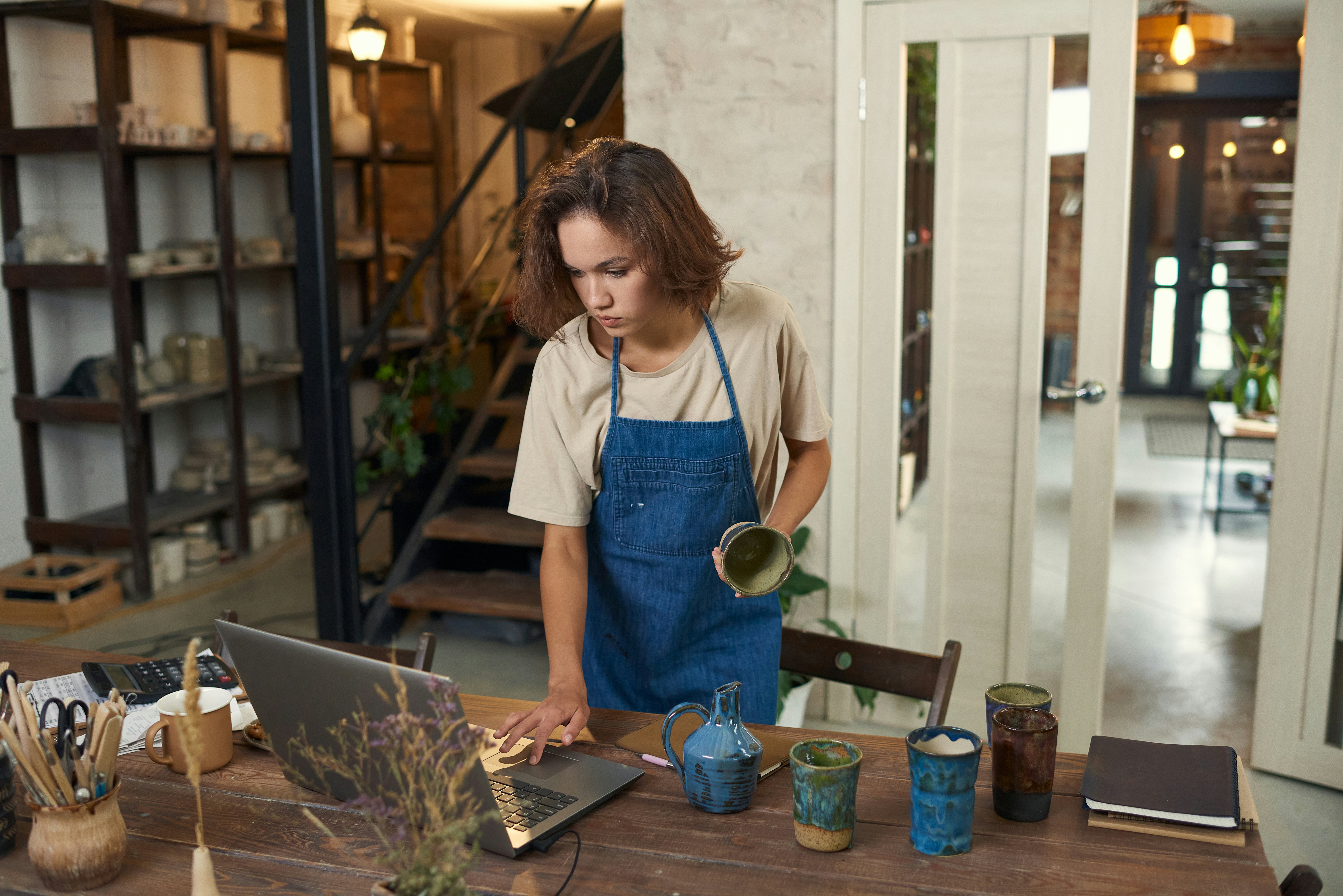 Woman in denim apron working at wooden desk with laptop and ceramic vessels in modern studio space.