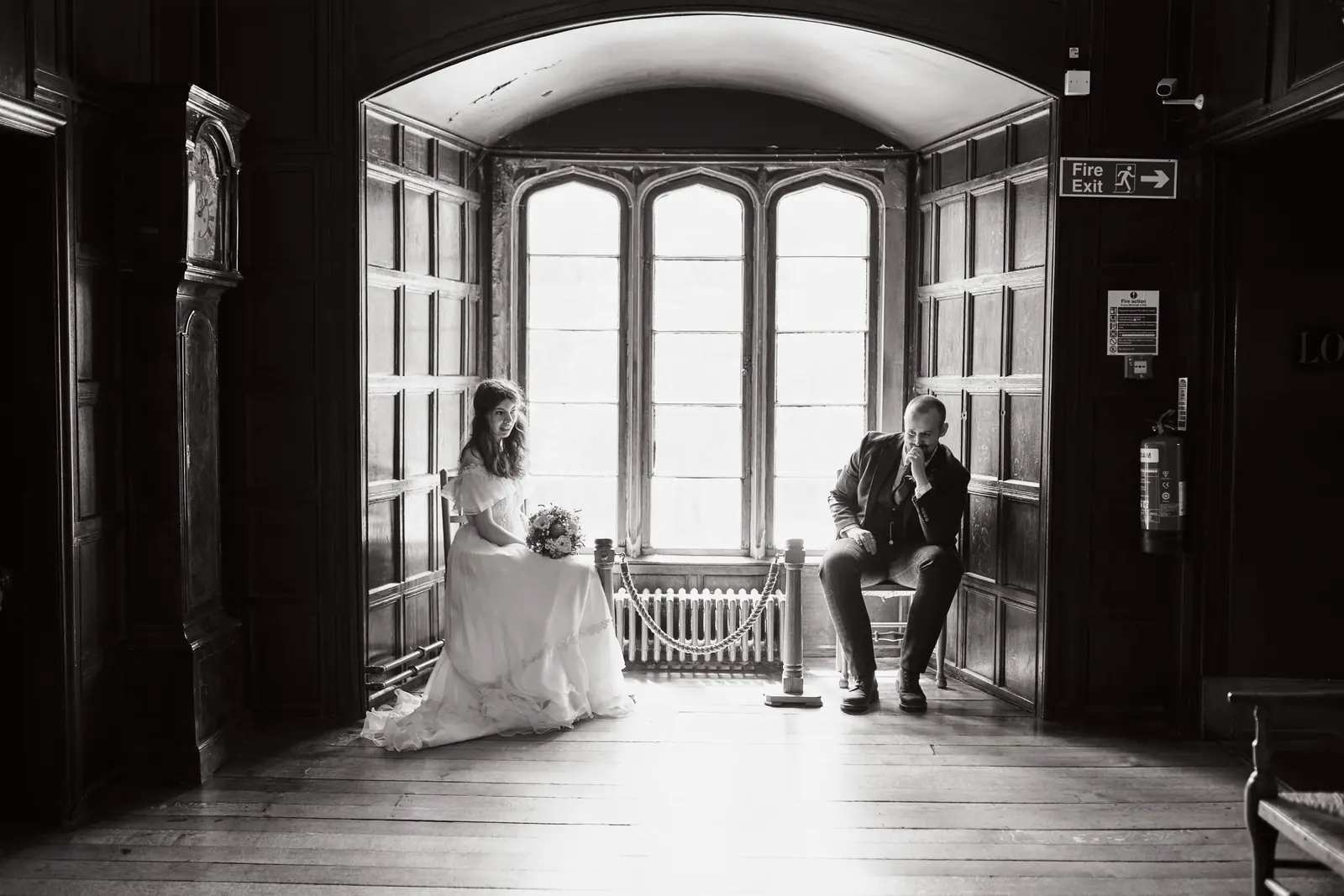 Black and white photo of a bride and groom sitting either side of a tall leaded window in a panelled room