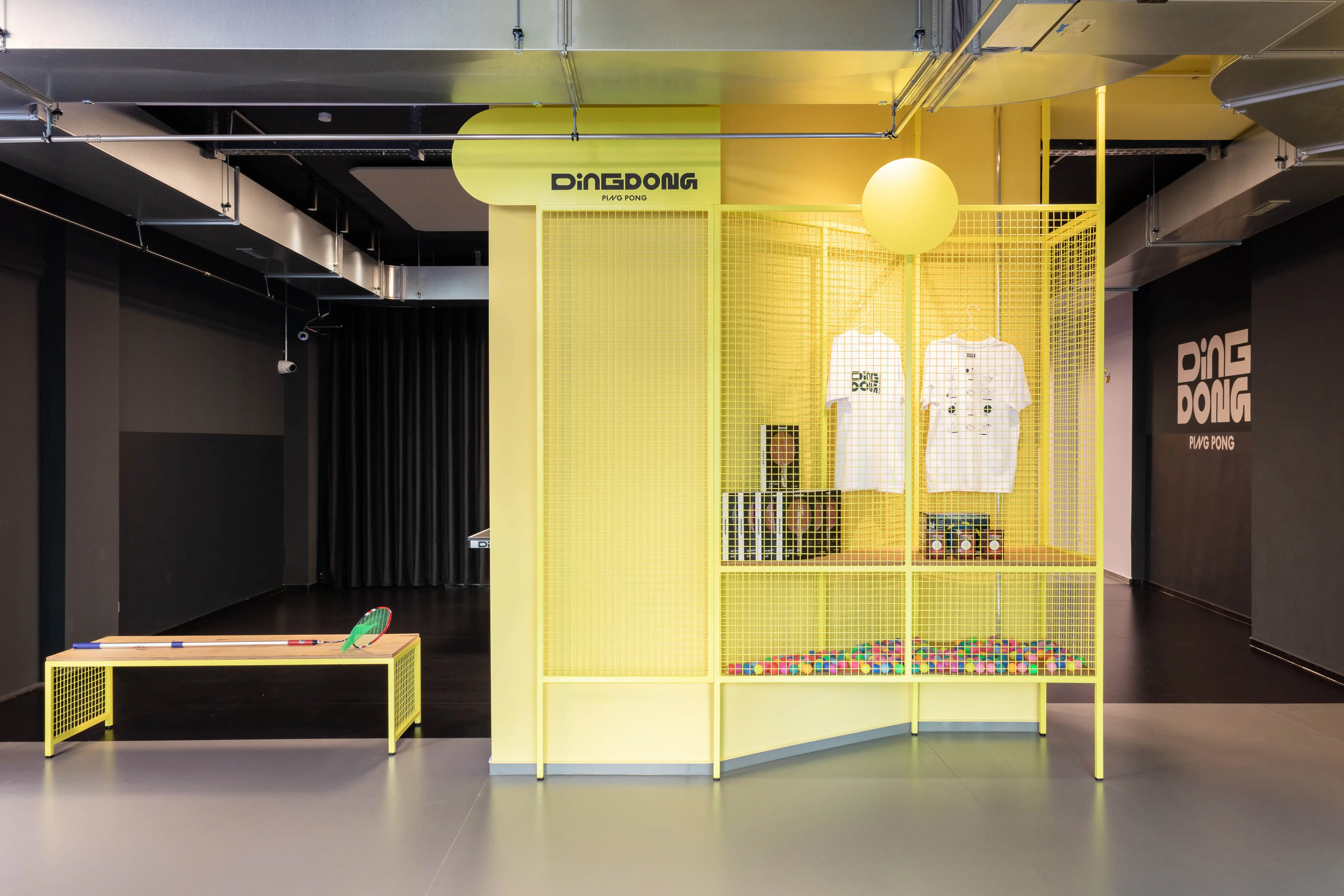Yellow wire-mesh merchandise display at DING DONG PING PONG with T-shirts, balls in a pit and a glowing sphere lamp, next to a yellow bench with paddles.