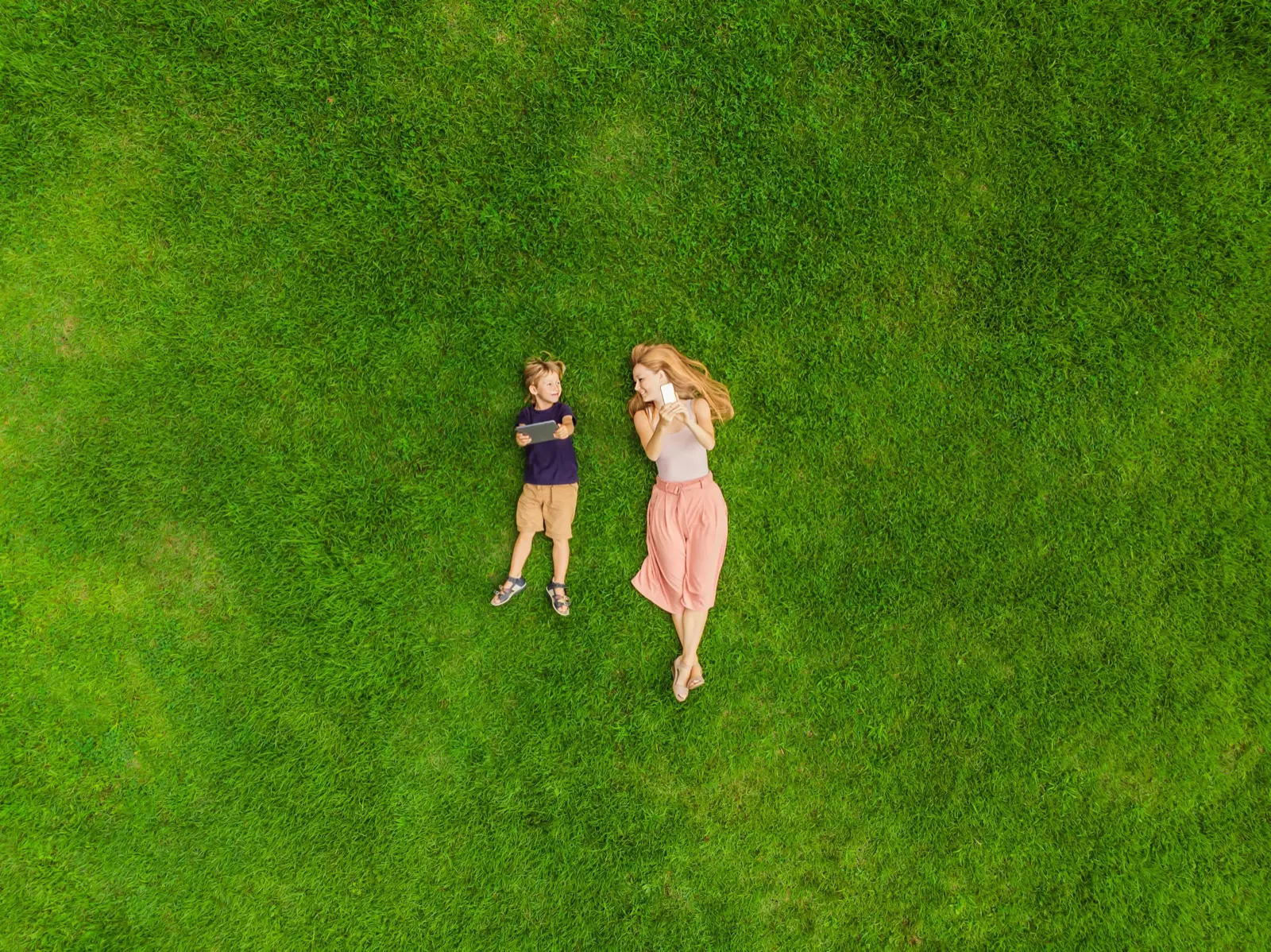 Aerial view of two people lying on green grass, one in a dark shirt and the other in a light dress.