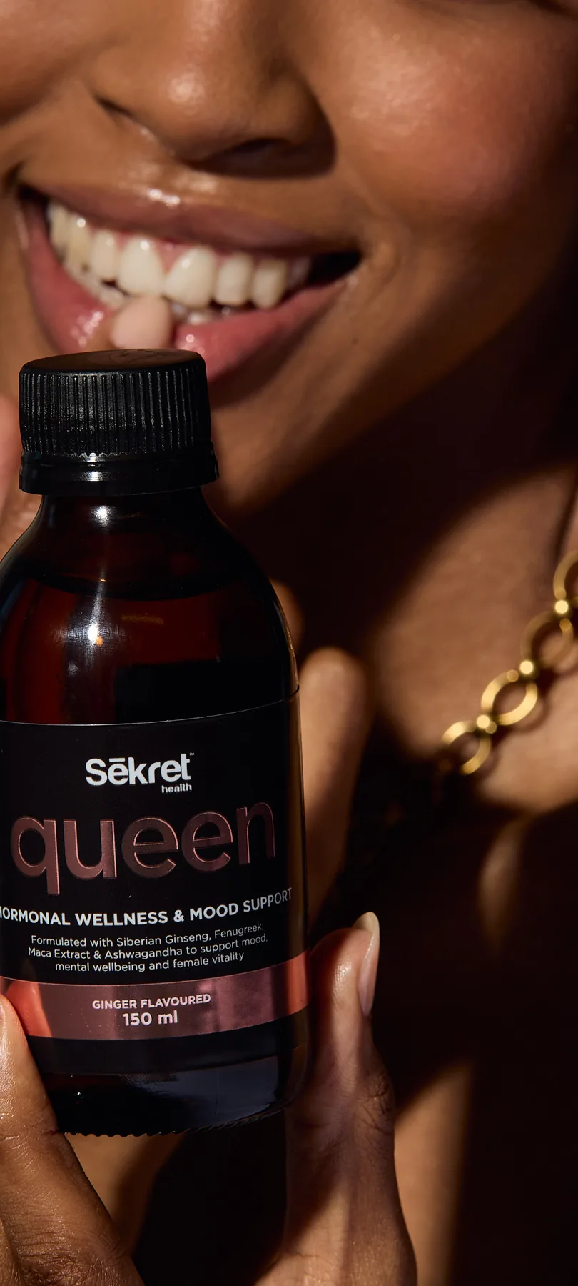 A smiling woman holds a bottle of Sēkret Queen syrup in golden light