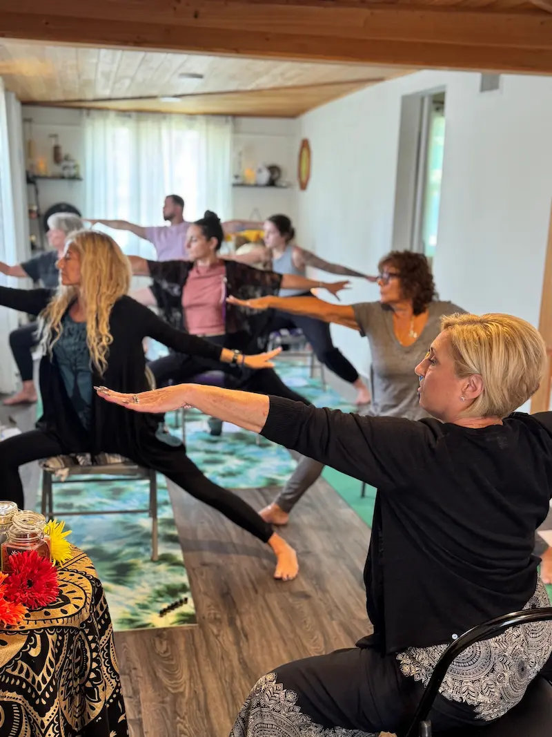 A group of people participate in a seated yoga class, each in a warrior pose while inside a cozy, light-filled studio in Bradenton, FL.