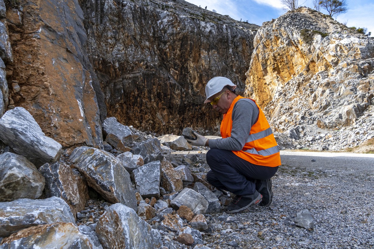 A worker in an orange safety vest examines rocks in a quarry, with rocky cliffs in the background.