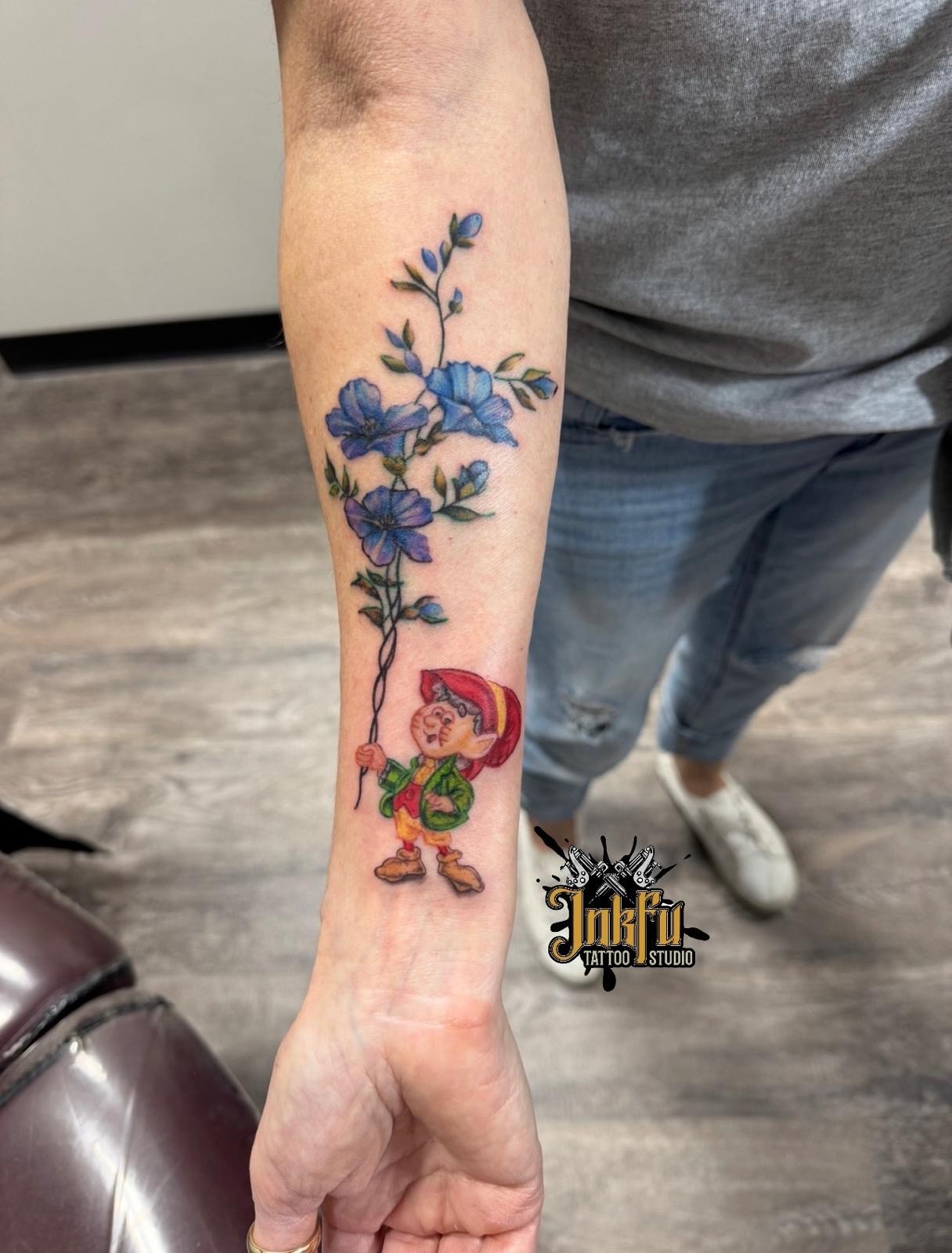 Color tattoo of blue flowers and a cartoon elf on the forearm, by Meer Rich