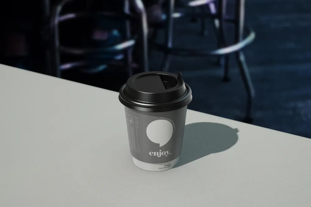 A black disposable coffee cup mockup with a matching lid, placed on a light gray surface. The minimal design features soft shadows, with a blurred cafe chair background adding depth.