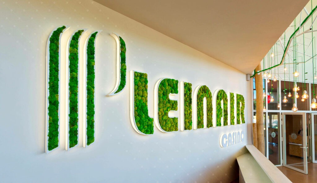 Enseigne Végétal fais par LeGraveur sur une murale avec l'inscription « PLEINAIR CASINO » en lettres lumineuses remplies de mousse verte. Le décor est moderne et lumineux, avec des suspensions décoratives.