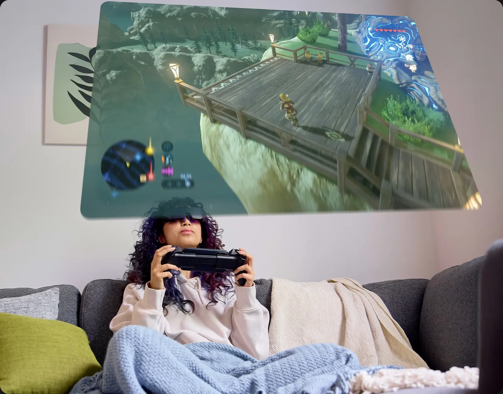 A person with curly hair sits on a couch, wrapped in a blue blanket, using a virtual reality headset and controller to engage with a fantasy adventure video game projected above them, where a character explores a wooden platform in a lush, green environment.