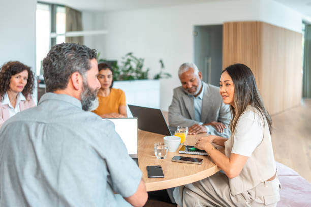 Two businessmen sitting at a table discussing work while looking at a laptop, in a bright office with natural light.