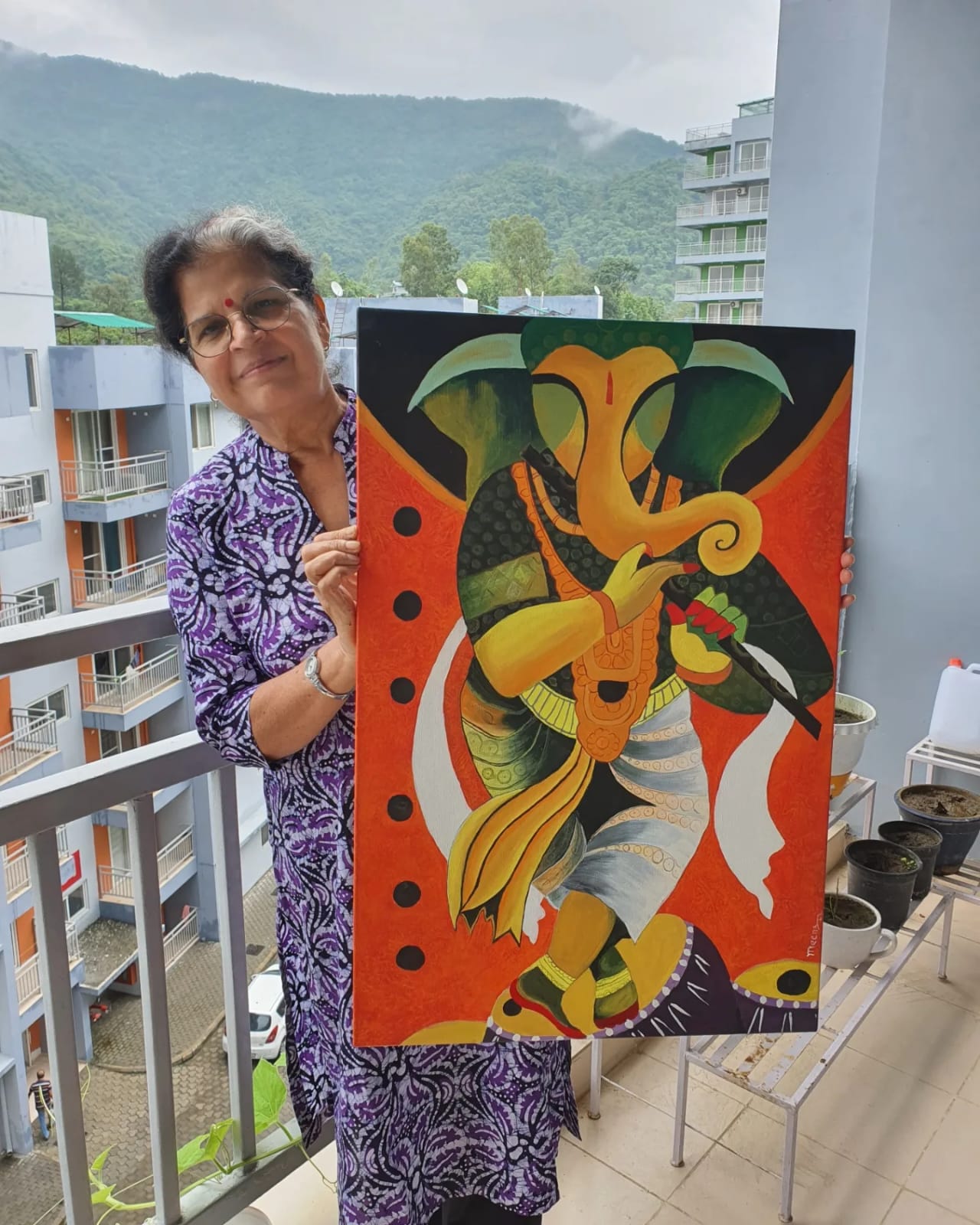 Creative Pod student holding a vibrant painting of Hindu God Ganesh playing a flute on the balcony of The Creative Pod studio.