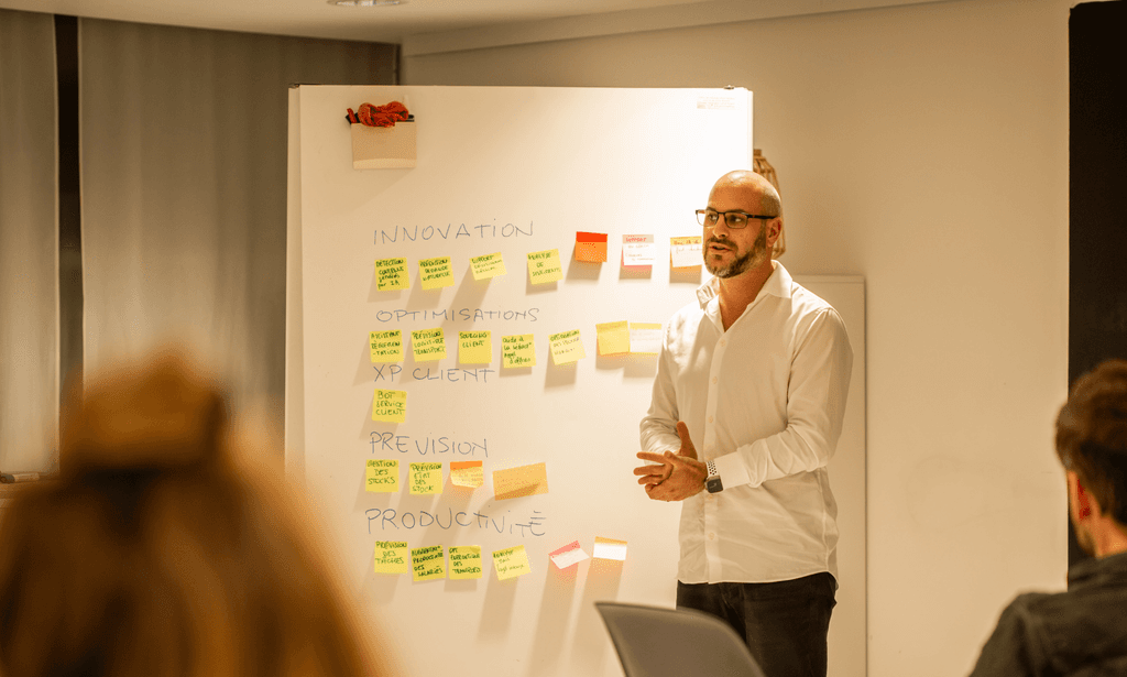 Homme en chemise blanche qui présente devant un tableau avec des post-it colorés face à une audience assise l’écoutant.