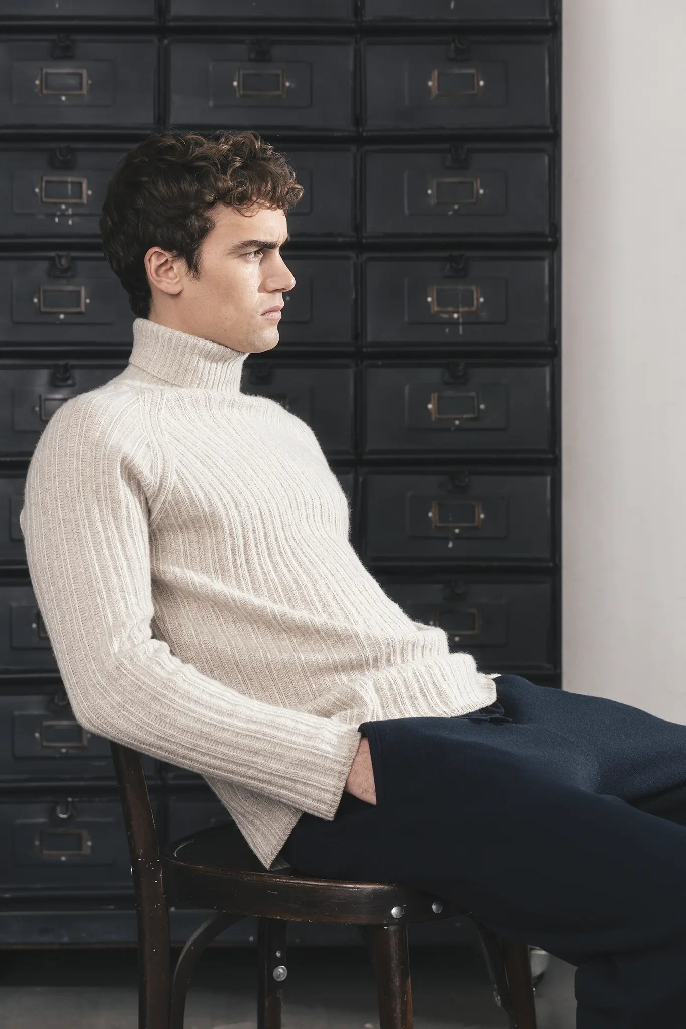Man seated on a wooden chair wearing ivory ribbed cashmere turtleneck and navy knit trousers – Aida Barni FW25/26.