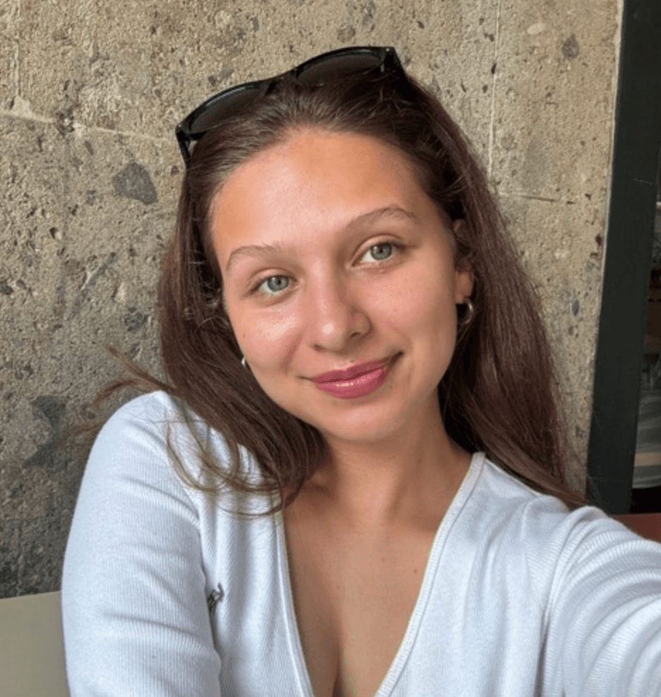 A person with curly hair and glasses smiles at the camera, seated in a cozy workspace with warm lighting.