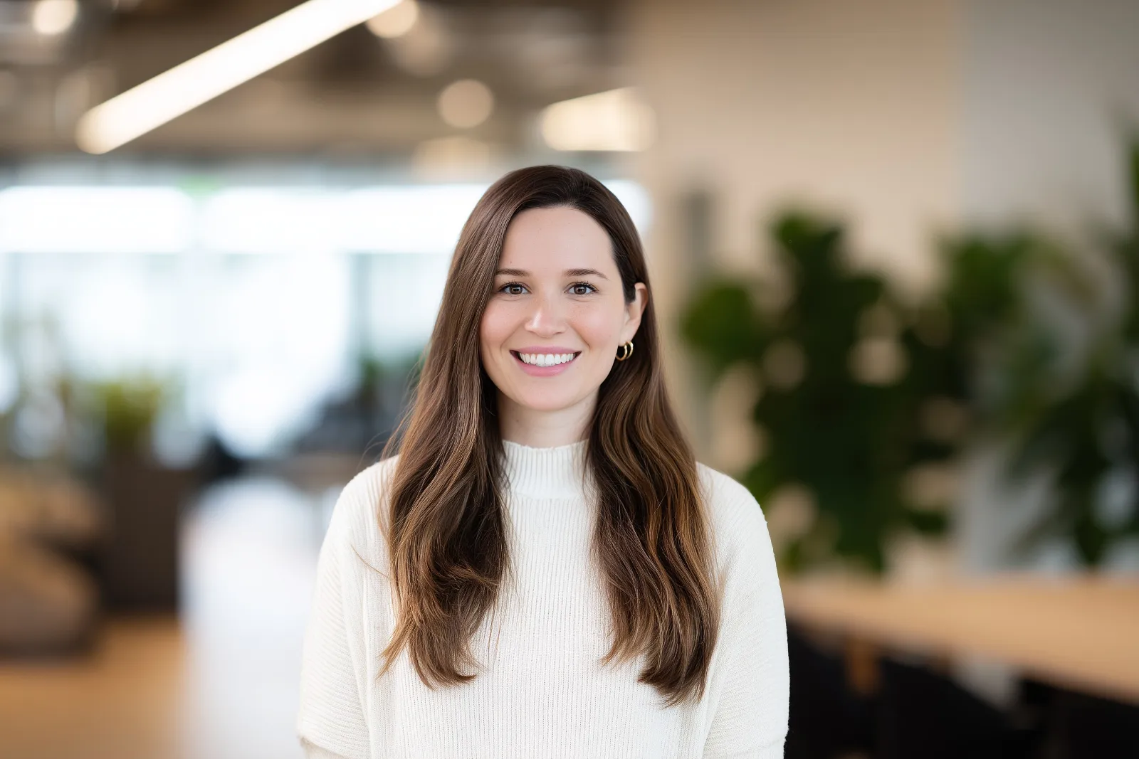 A woman with long brown hair wearing a white knit sweater stands in a bright office.