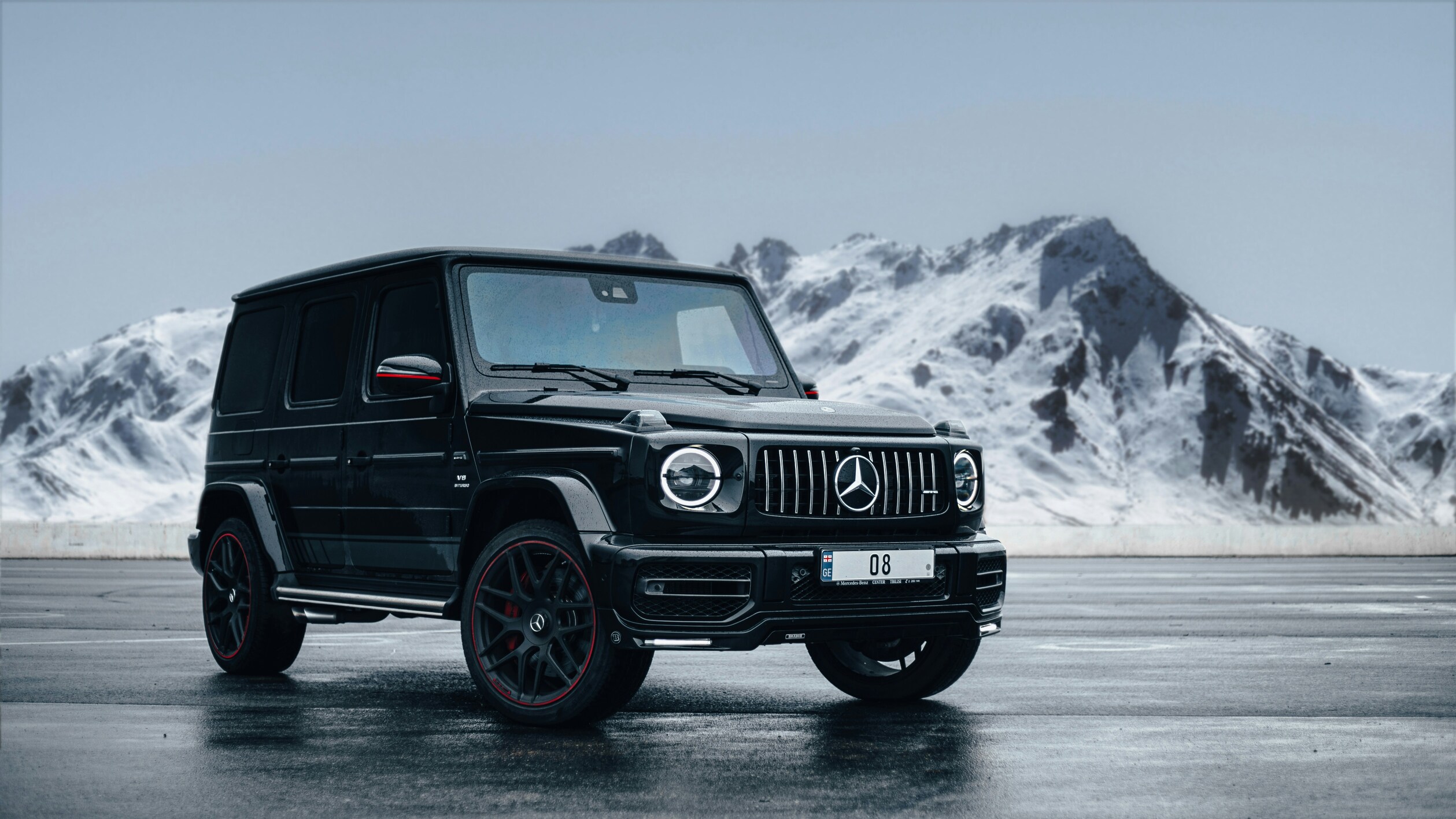 A sleek black Mercedes-Benz G-Class SUV is parked on a wet roadway against a backdrop of snow-covered mountains, showcasing its rugged elegance and luxury.