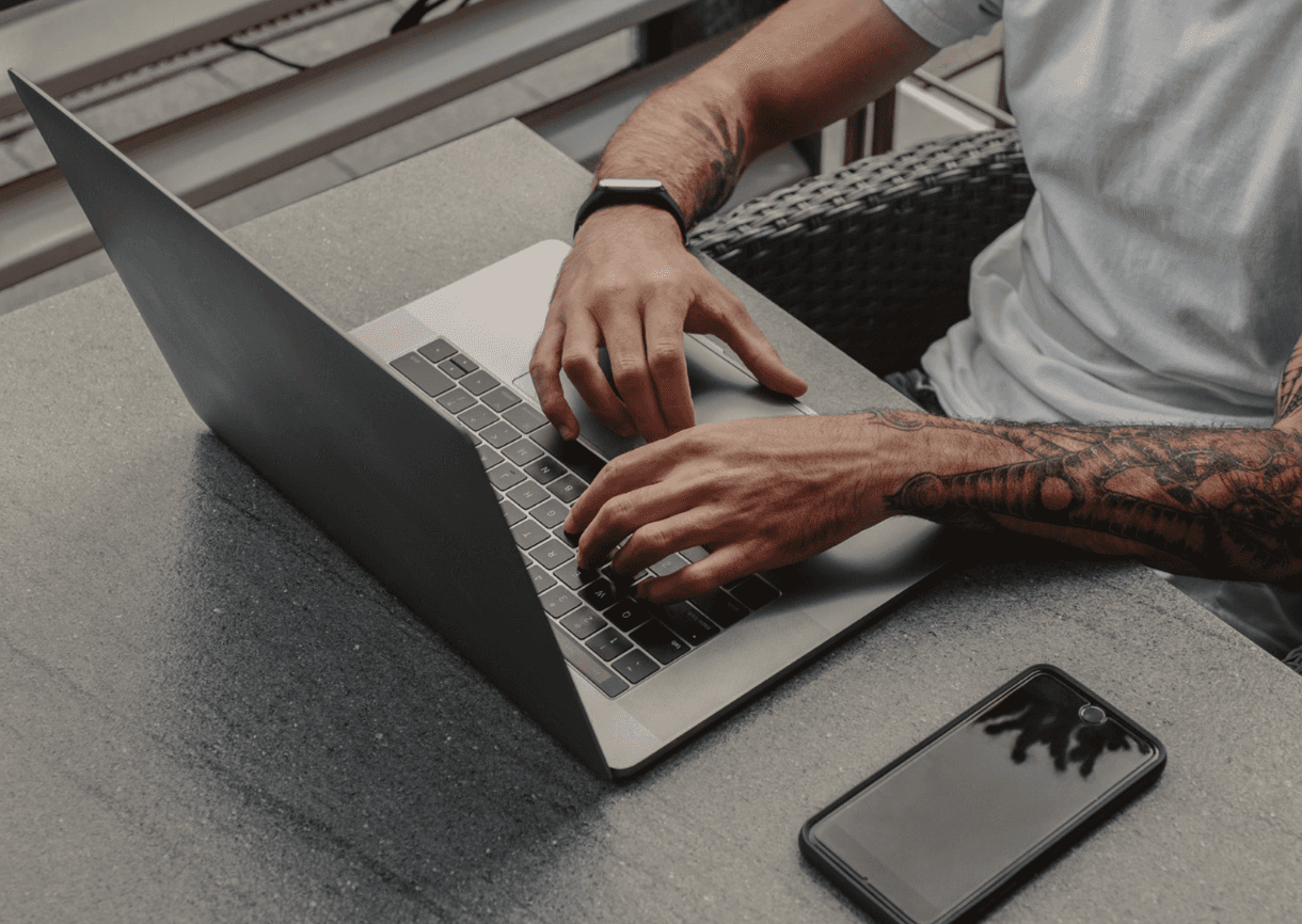 A man in a brown blazer and a woman in a light sweater sit at a desk with a laptop, discussing something. The room is dimly lit, creating a focused atmosphere.