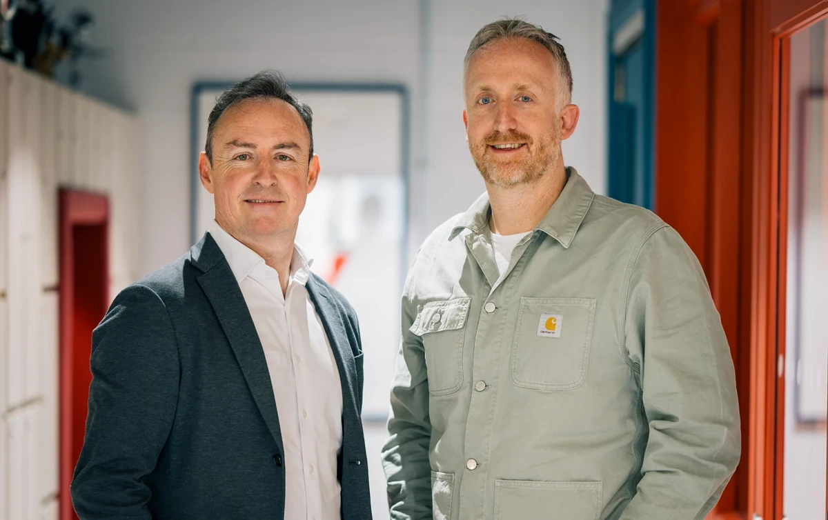 Two men standing side by side in a corridor with red and white walls. they are looking at the camera and smiling