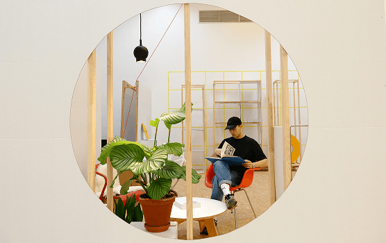 Blick durch ein rundes Öffnungselement einer Regalwand auf ein Sitzensemble, in dem eine Person ein Magazin liest; im Hintergrund Regale und Wandgrid des modularen Store-Konzepts.