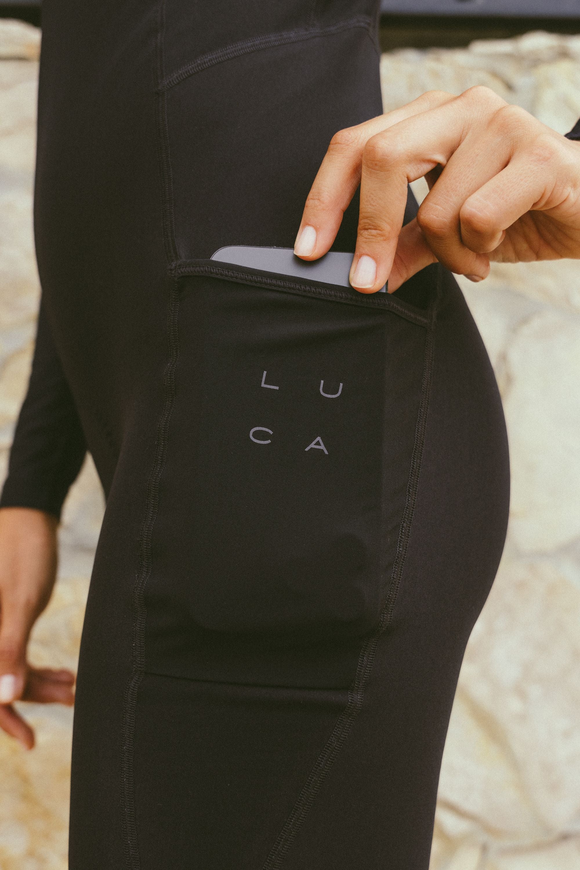 Close-up-of-black-athletic-Stride-Bib-Tight-highlighting-a-deep-side-pocket- designed-to-hold-a-cell-phone-A-hand-is-inserting-a-dark-colored-phone-into-the-pocket-The- pocket-area-features-a-subtle-matte-gray-LUCA-logo-printed- vertically-arranged-in-two-rows-of- two-letters-The-background-is-a- blurred-textured-stone-wall