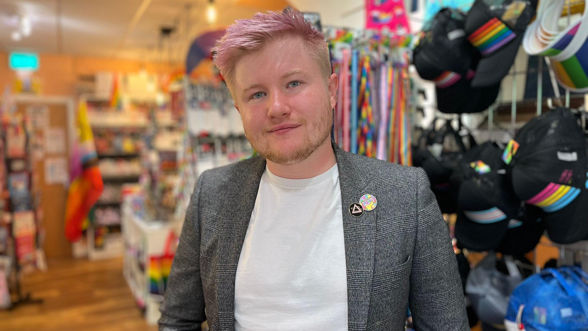 A man wearing a white t-shirt and a grey suit jacket. He has stubble, blue eyes and light pink hair. In the background are shop shelves, many items have rainbow emblems. 