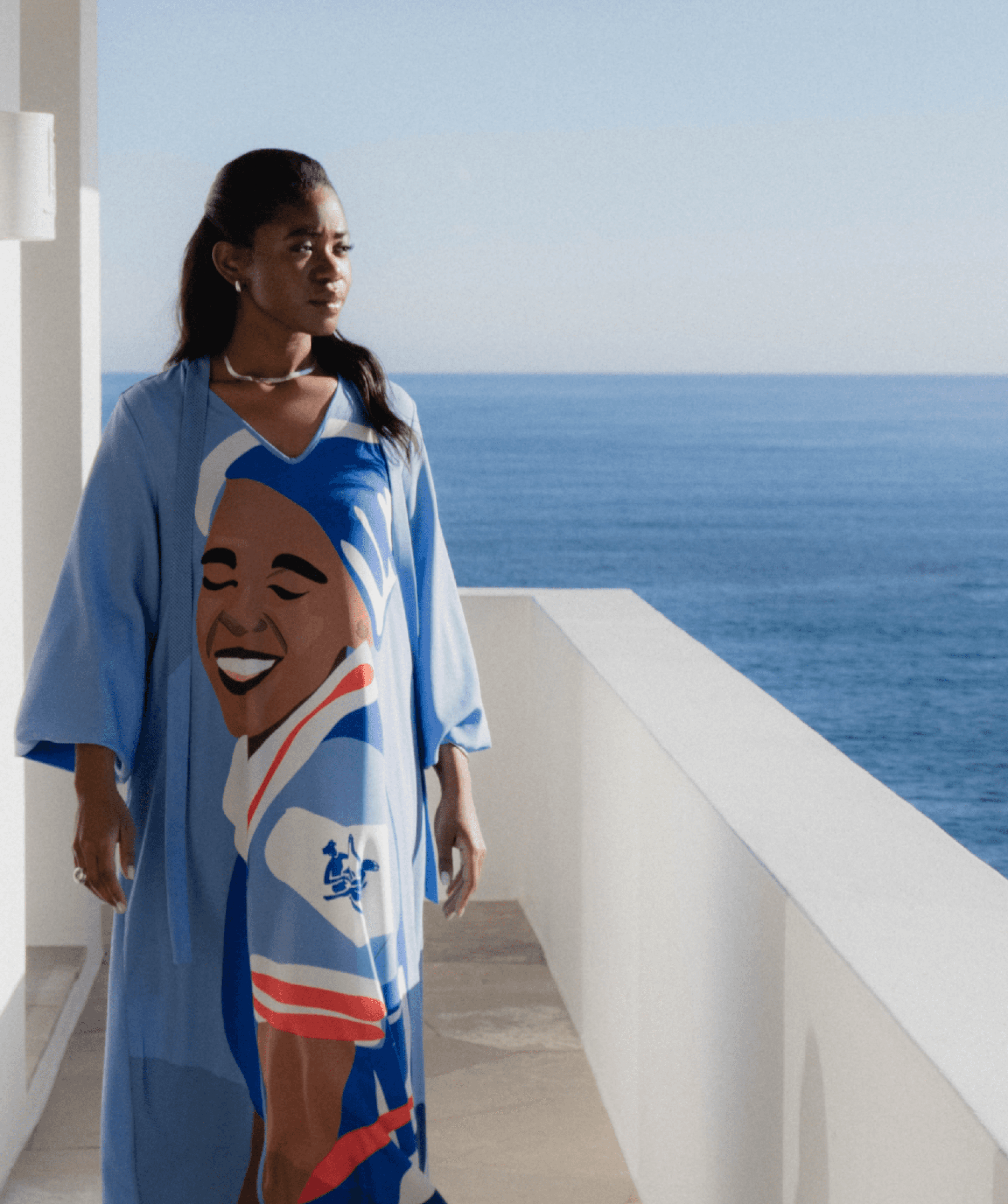 Vimbai in a blue printed kaftan dress standing on a white oceanfront terrace with a thatched wooden ceiling and sea view