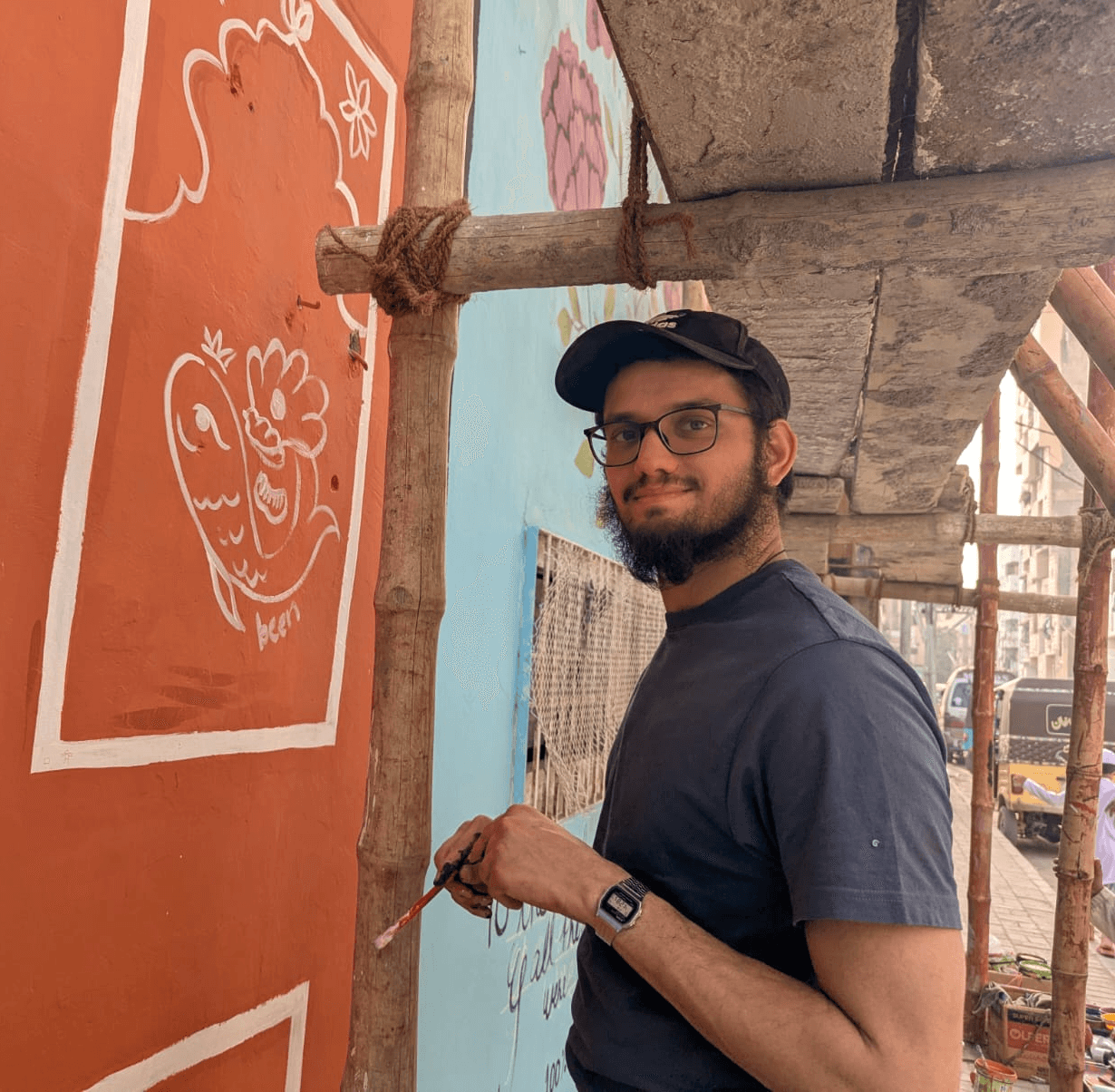 picture of ceo shabbir muhammad looking at the camera, wearing a cap, glasses in a grey shirt while holding a paintbrush infront of an orange and white mural