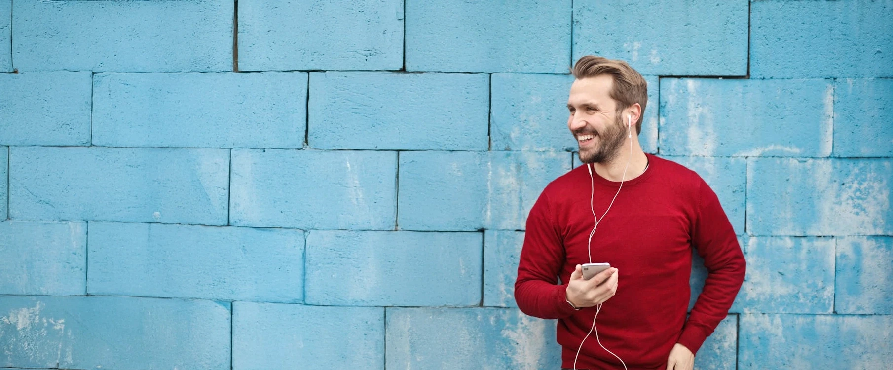 A man similing at his phone leaning against a blue brick wall