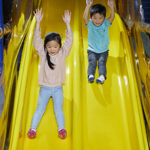 Dvoje djece spušta se niz jarko žuti tobogan, oboje s podignutim rukama i izrazima oduševljenja na licima.