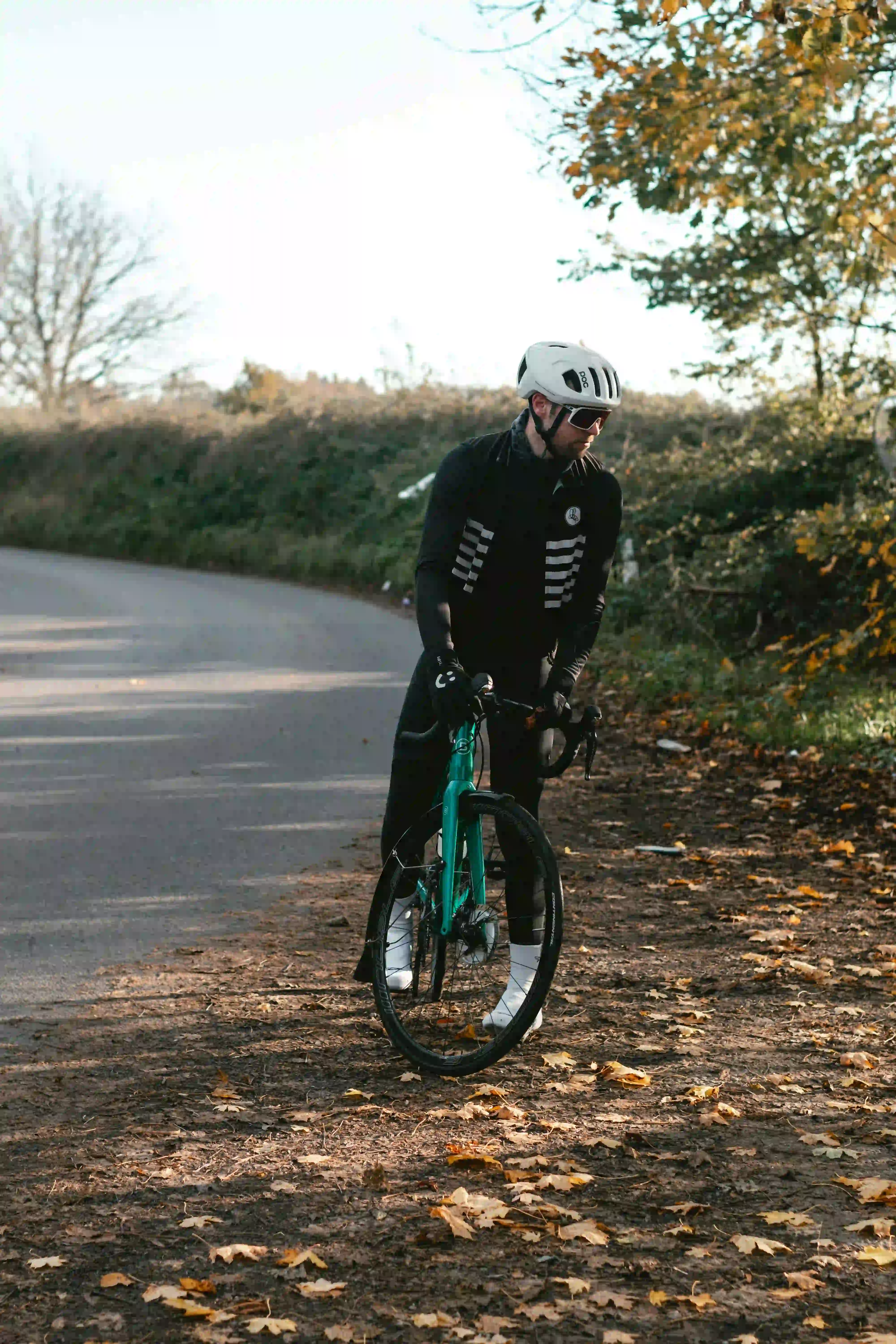 Cyclist standing with a road bike on a leaf-covered path wearing a helmet and black cycling gear.
