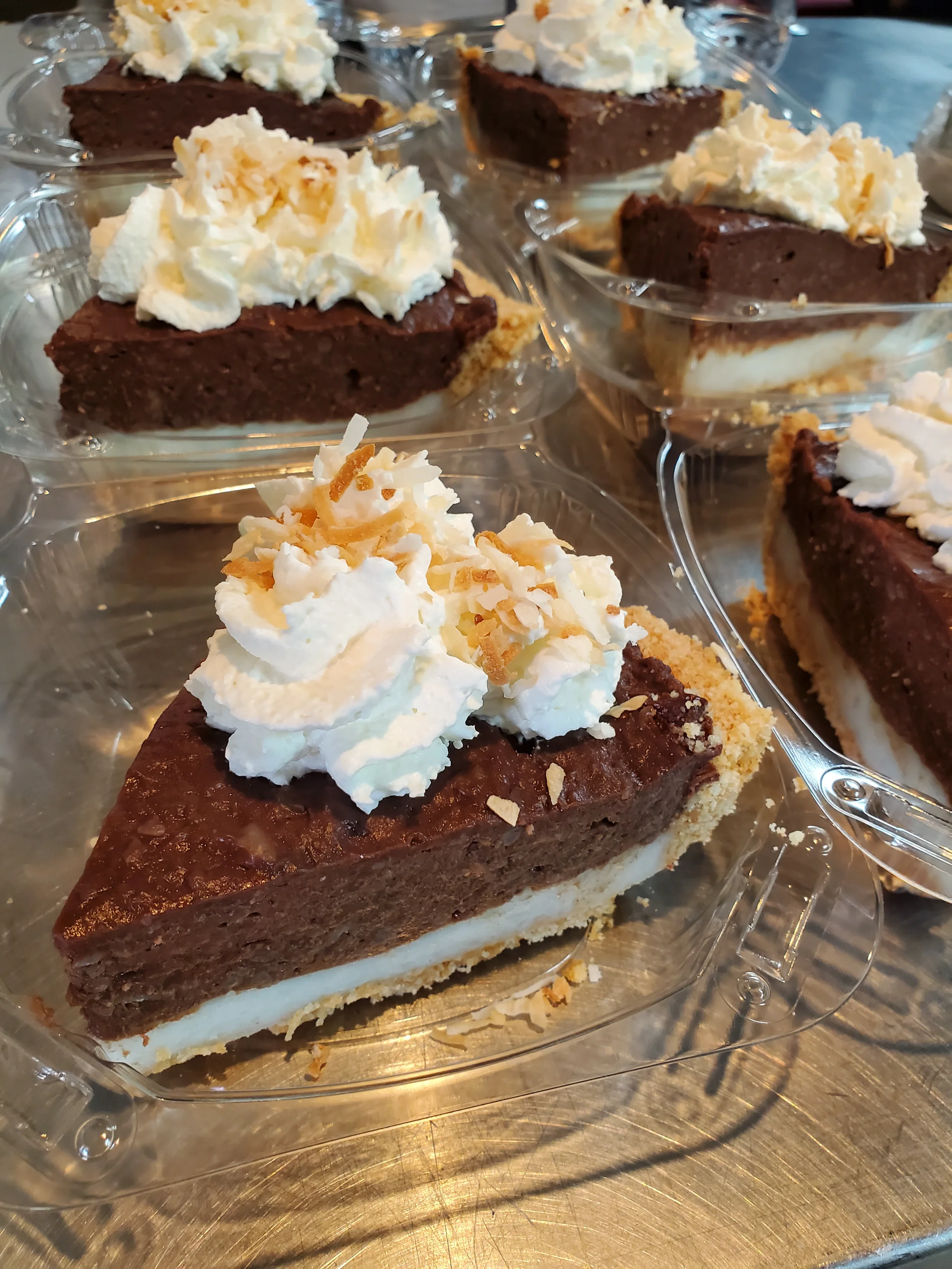 Multiple slices of Chocolate Haupia Pie in clear containers, showing distinct layers of dark chocolate and white coconut haupia topped with whipped cream and toasted coconut.