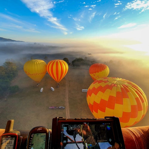 Verschillende luchtballonnen in levendige gele en oranje tinten zweven laag over een mistig veld bij zonsondergang, met navigatieapparatuur op de voorgrond.