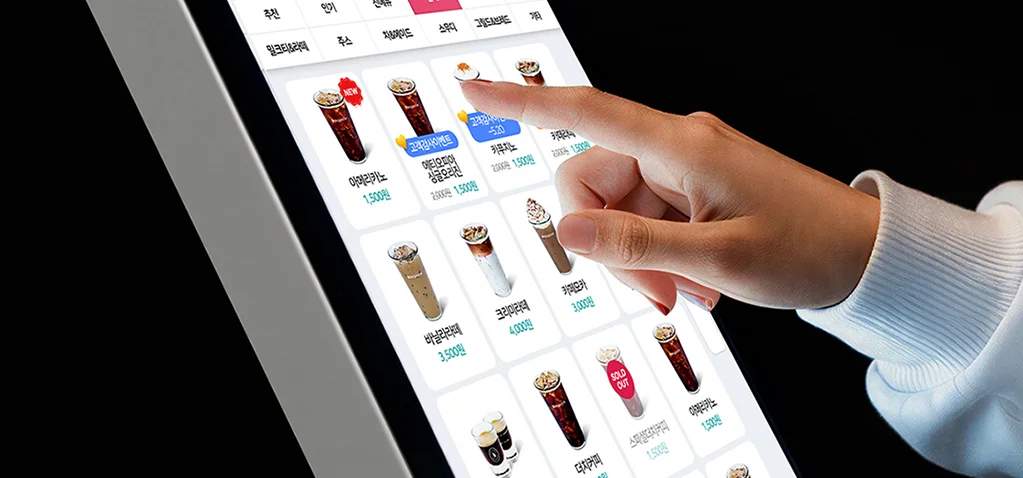 다양한 음료 메뉴가 배열된 키오스크 화면에서 항목을 터치해 선택하는 장면
