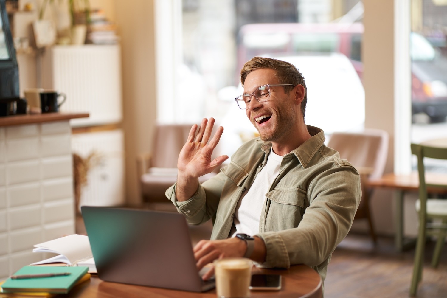 Junger Mann mit Brille sitzt an einem Tisch im Café vor einem Laptop und winkt mit der rechten Hand