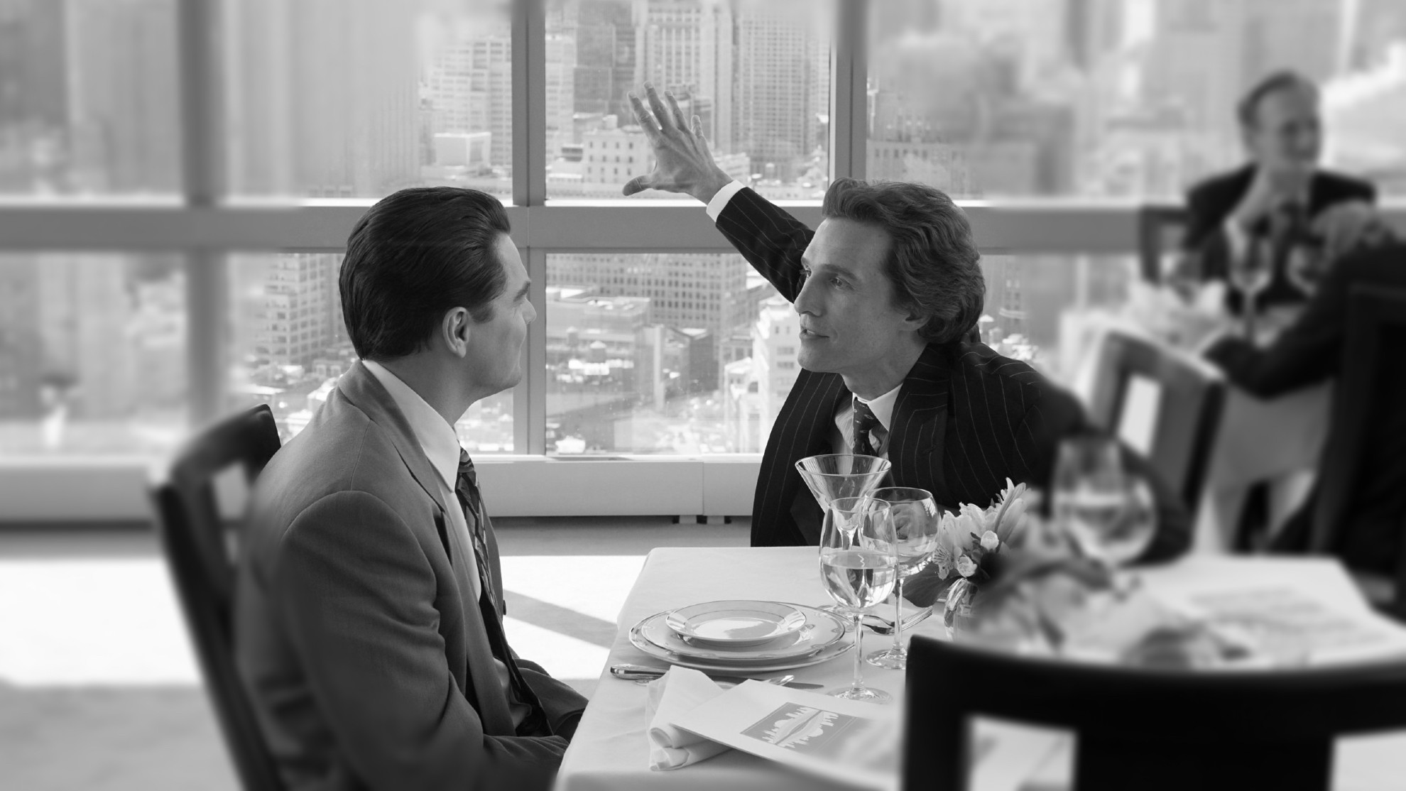 Black and white photo of 2 men sitting one in front of each other in a restaurant. On of them id dramatically explaining something to the other one. on the background you see New York's skyscrapers