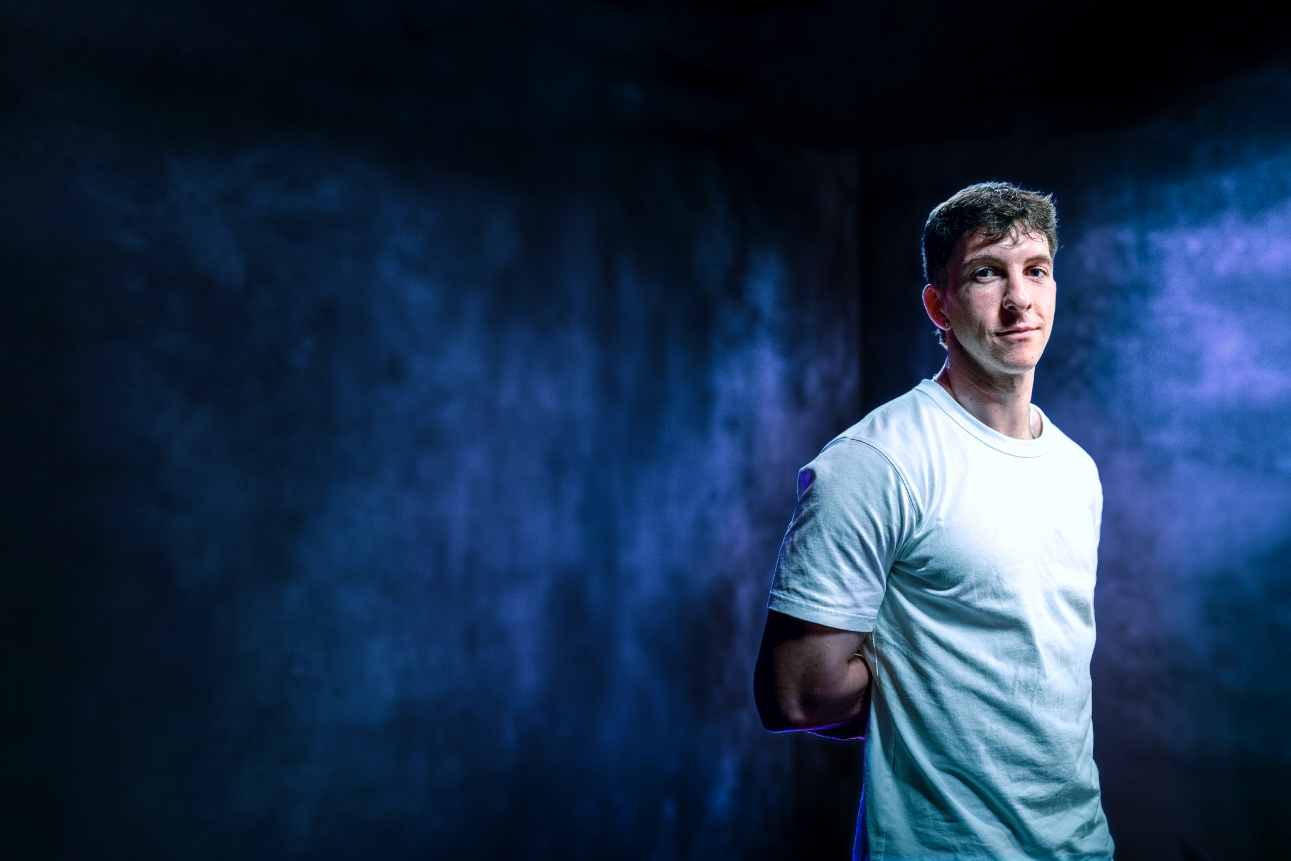 Portrait professionnel et moderne d'un jeune homme, représentant le fondateur ou directeur artistique de Nux-Studio, posant de face avec un léger sourire. Il porte un simple t-shirt blanc à manches courtes. L'arrière-plan est un mur texturé, sombre et baigné d'un éclairage dramatique de couleur bleu électrique et violet, créant une ambiance de studio créatif et innovante. L'espace négatif important sur la gauche est idéal pour l'ajout d'un titre, d'un logo, ou d'un slogan. Ce visuel évoque l'expertise en branding, direction artistique et l'identité moderne d'une agence de design digital.