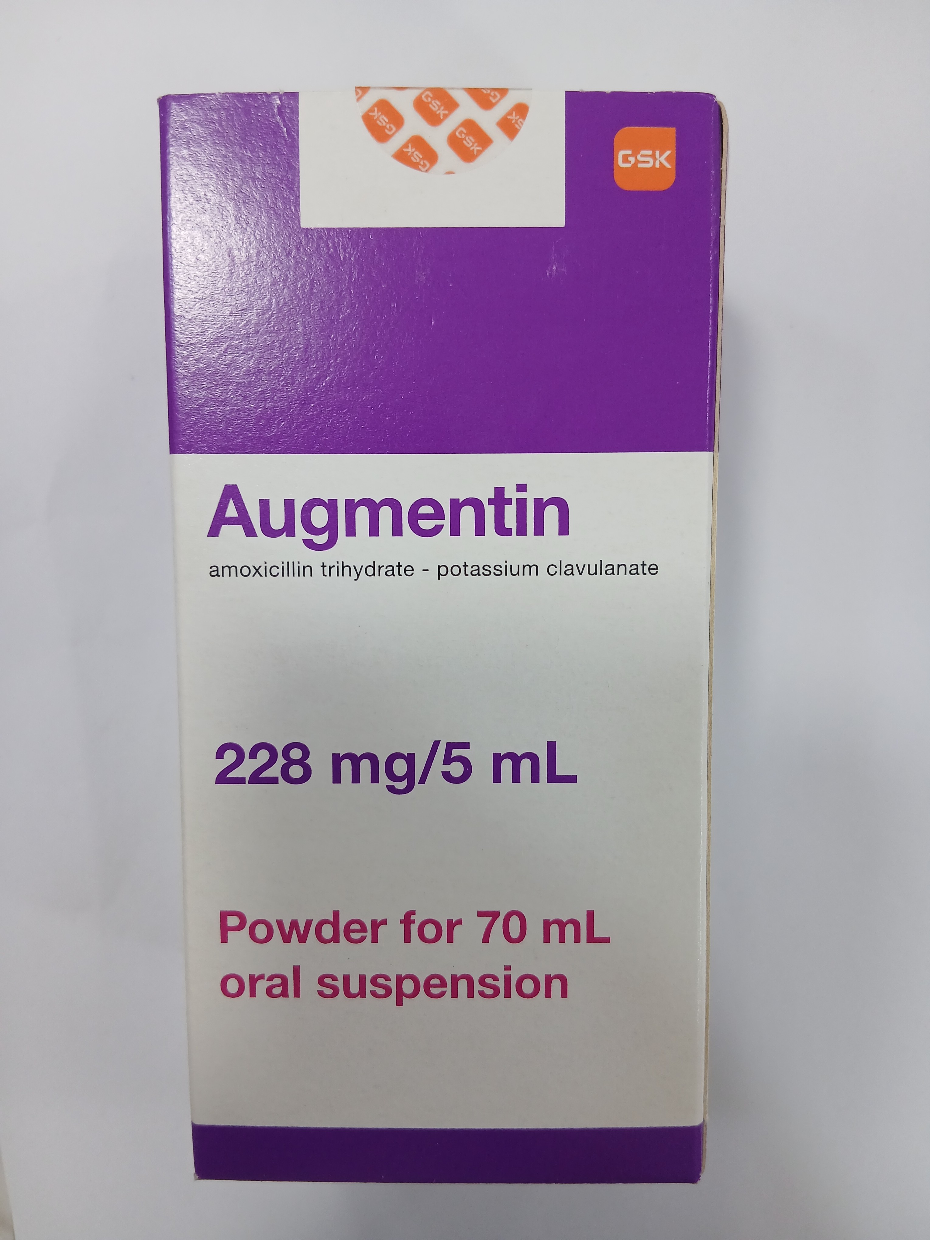 A white and purple cardboard box for Augmentin oral suspension is shown, displaying "228 mg/5 mL" and "Powder for 70 mL oral suspension" in purple text, with an orange GSK logo in the top right corner.