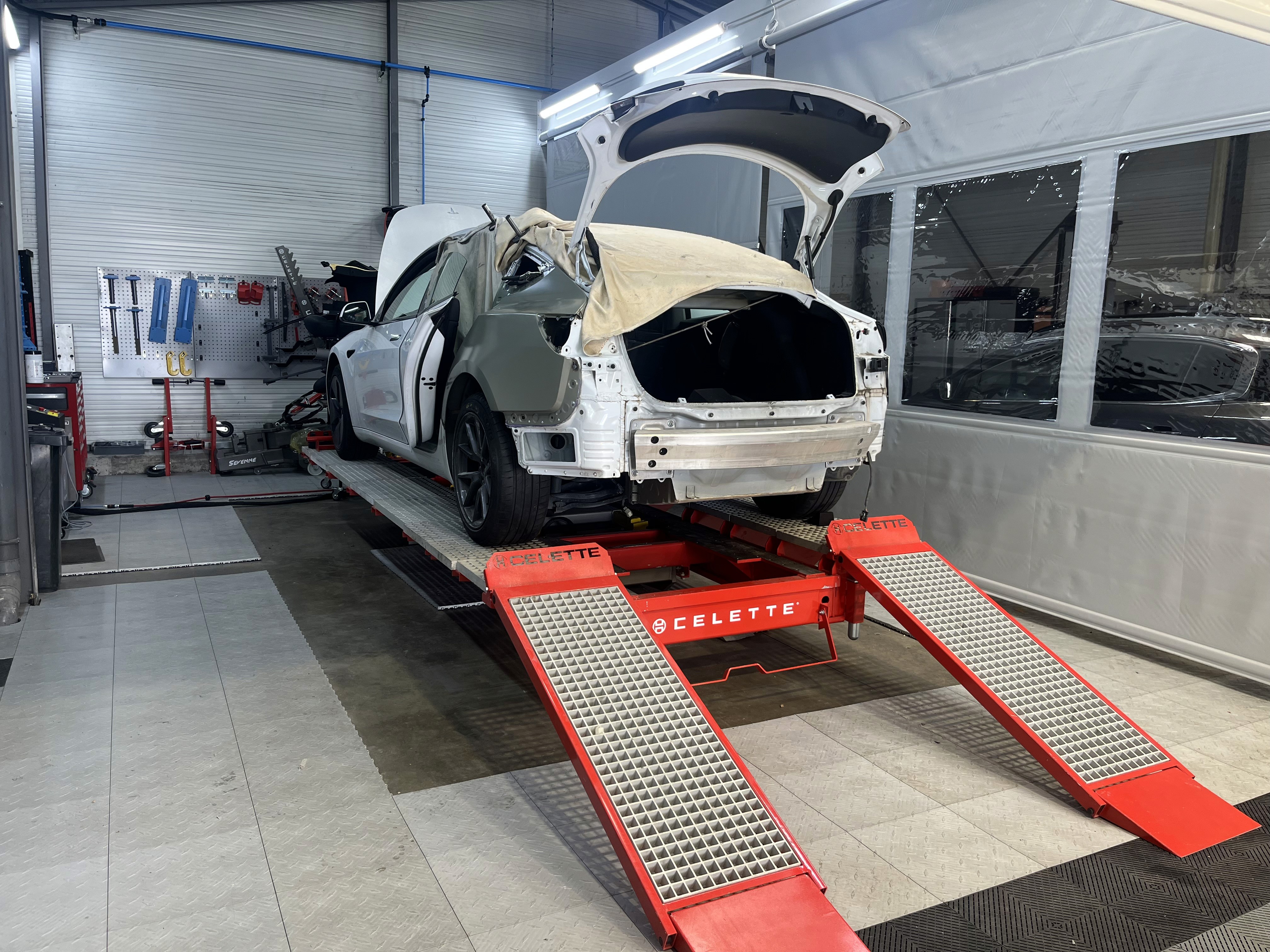 Two people in a workshop, one standing and smiling while the other leans over a car, working on it.