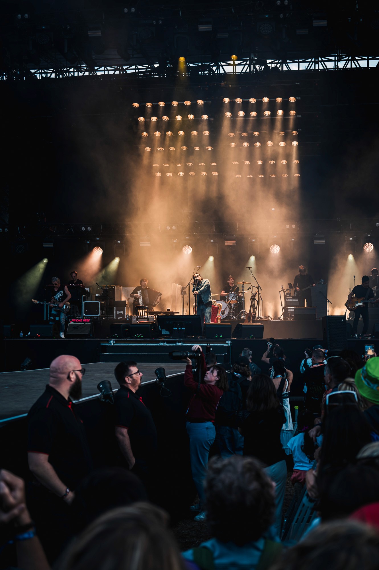 Groupe sur scène dans une brume dorée, sécurité au premier plan, concert de Saez