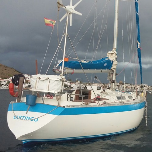 A sailboat named "Vartingo" docked in a harbor, under a cloudy sky, with a town and hillside in the background.