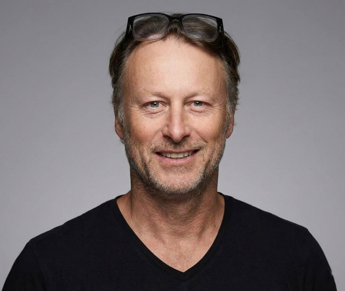 A smiling man with short hair stands with arms crossed, wearing a dark shirt against a neutral background.