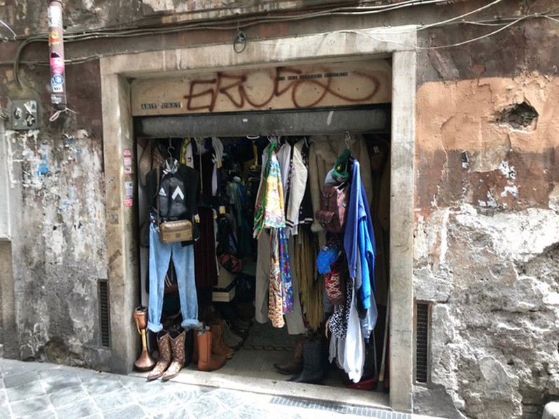 A photo of a store's entrance, made from stone, where there are cloths hanging and some shoes on the sides.