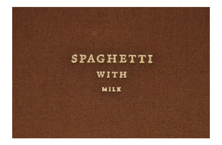 A dark brown notebook with a black elastic band, featuring the title "SPAGHETTI WITH MILK" printed in light gray, stands upright on a light beige surface.